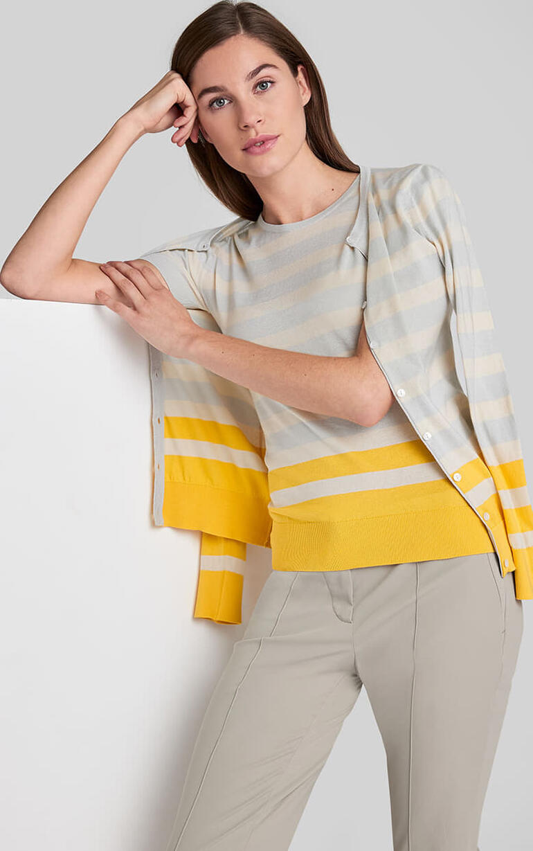 Femme portant un twinset rayé Smedley en jaune et écru. Elle s'appuie contre un mur.