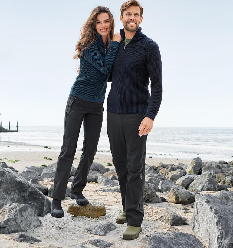 Couple en pulls marins sur la plage, entourés de pierres et de la mer.