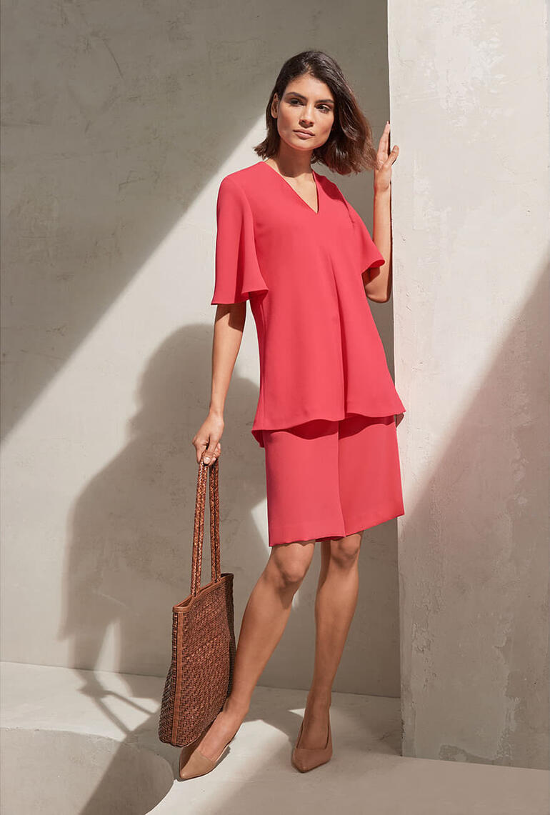 Chemisier col en V et Bermuda Femme en haut et jupe rose souriante devant un mur