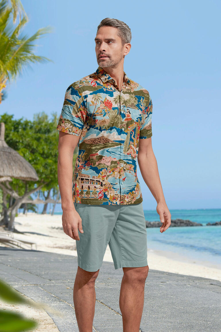 Homme en chemise Aloha colorée se tenant sur la plage, entouré de palmiers.