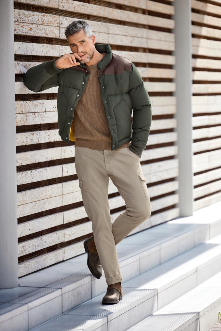 Veste Ranger Schott NYC Homme portant un pull en laine de chameau et une veste, assis sur des rochers.
