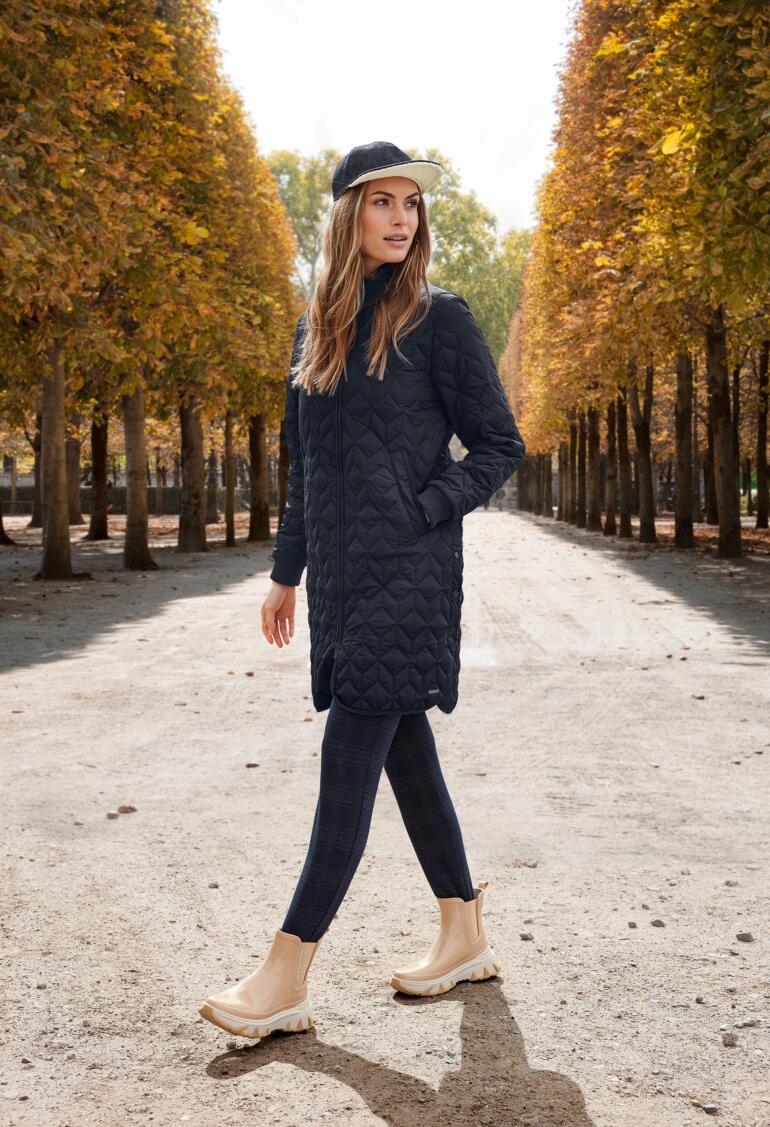 Femme en manteau matelassé noir marchant sur un chemin bordé d'arbres.