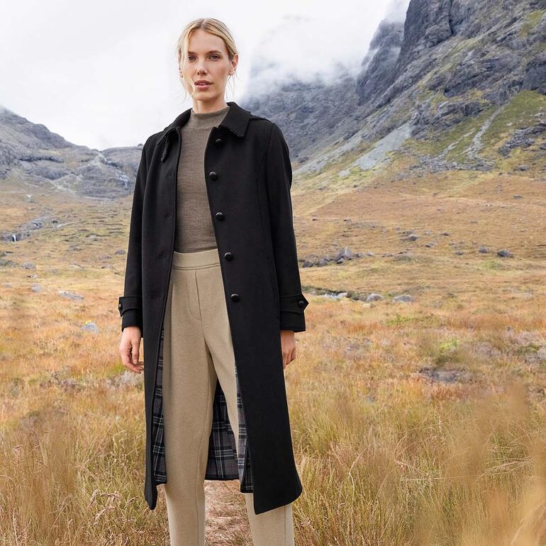 Beige Manteau en loden alpaga noir de Steinbock, porté par une femme dans la nature avec un pantalon clair.