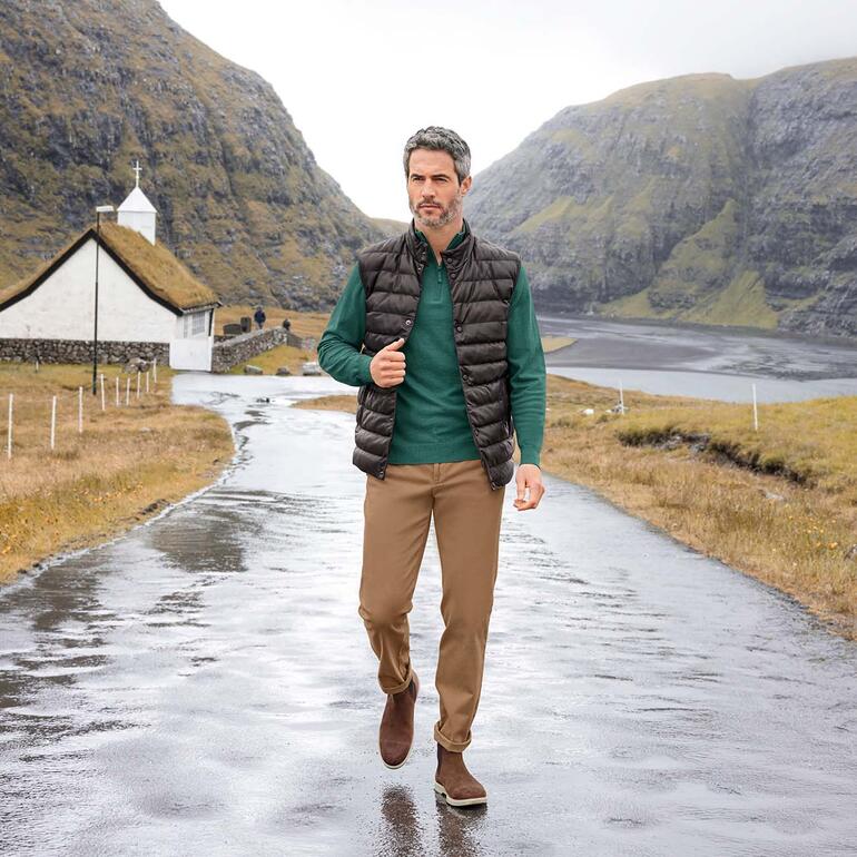Homme en pull toute saison vert et gilet noir marchant sur une route.