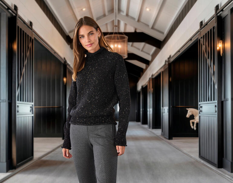 Femme en pull noir et pantalon pied-de-poule dans un hall avec un cheval en arrière-plan.