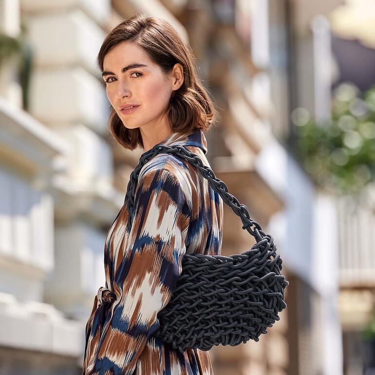 Femme avec un sac tressé dans une robe rayée se tient dehors.