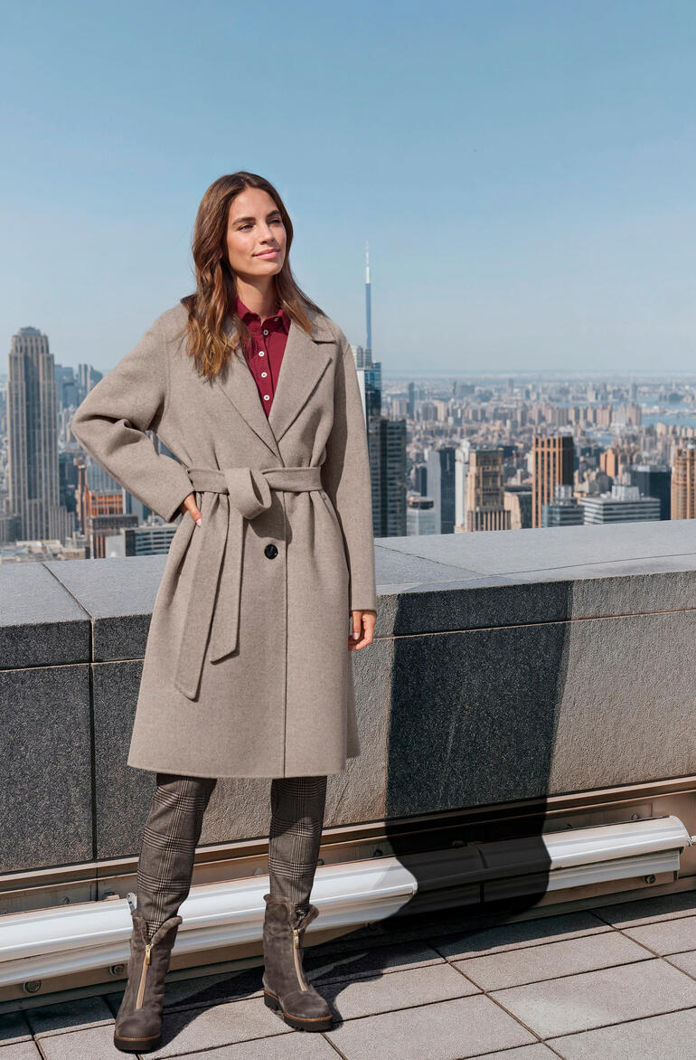 Femme aux cheveux bruns dans un manteau gris se tient sur un toit avec vue sur la ville.