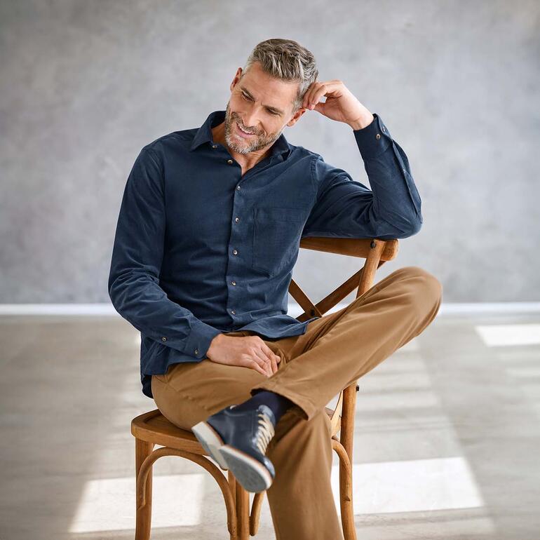Homme en chemise en velours côtelé bleu marine assis de manière décontractée sur une chaise.
