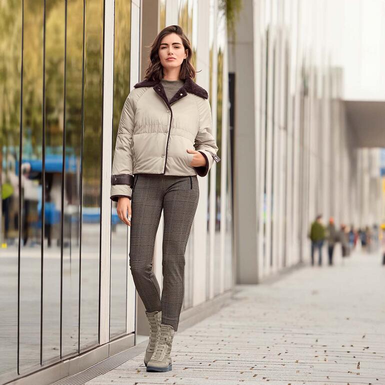 Femme en veste réversible en peau de mouton beige devant des bâtiments modernes.