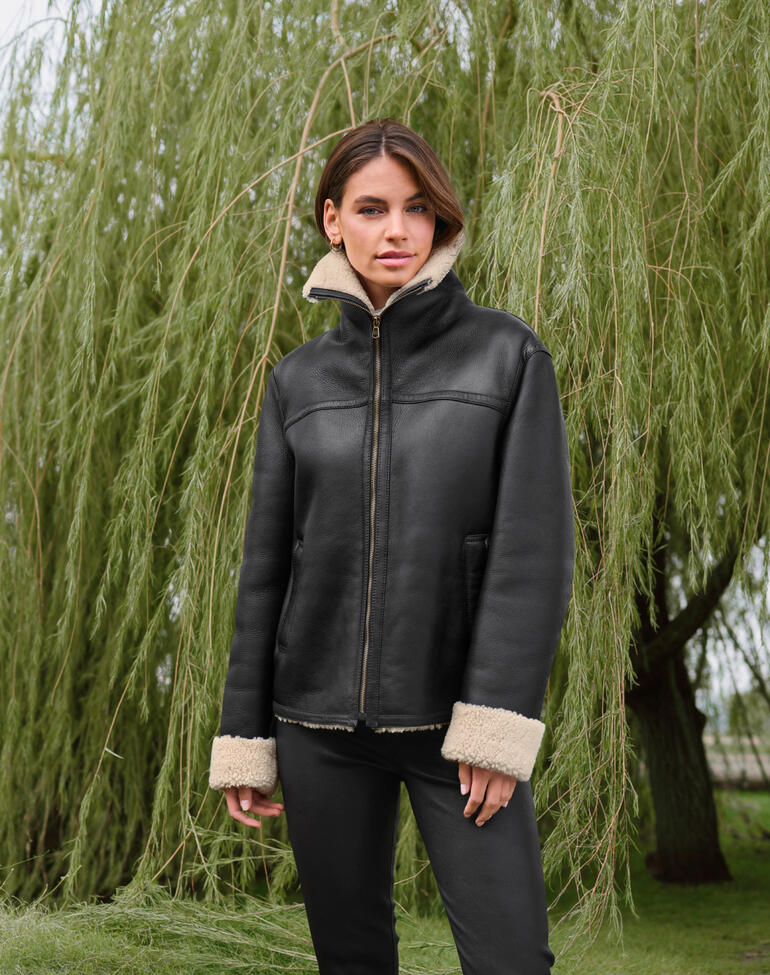 Femme portant une veste pilote en agneau bouclé noire devant un saule vert.