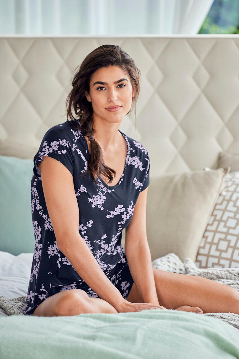 Femme en pyjama noire à motifs floraux souriante sur le lit.