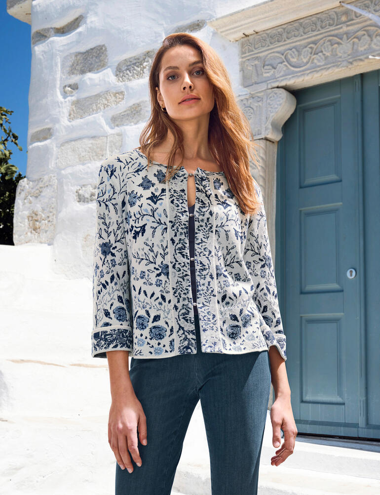 Femme portant un cardigan fleuri devant un mur blanc.