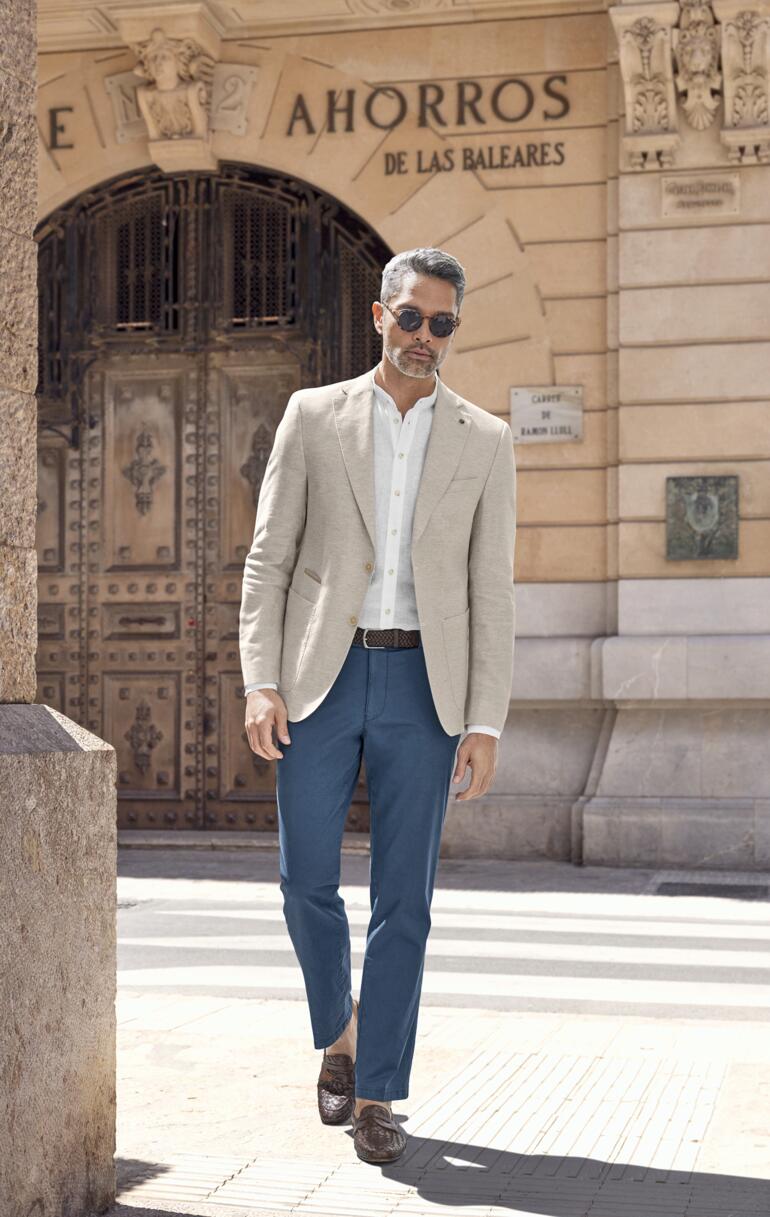 Homme en vest en lin beige business, portant un pantalon bleu, marchant devant un décor historique.