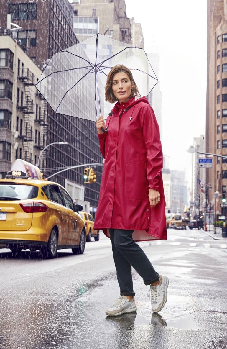 Femme en manteau de pluie rouge tenant un parapluie transparent dans un environnement urbain.