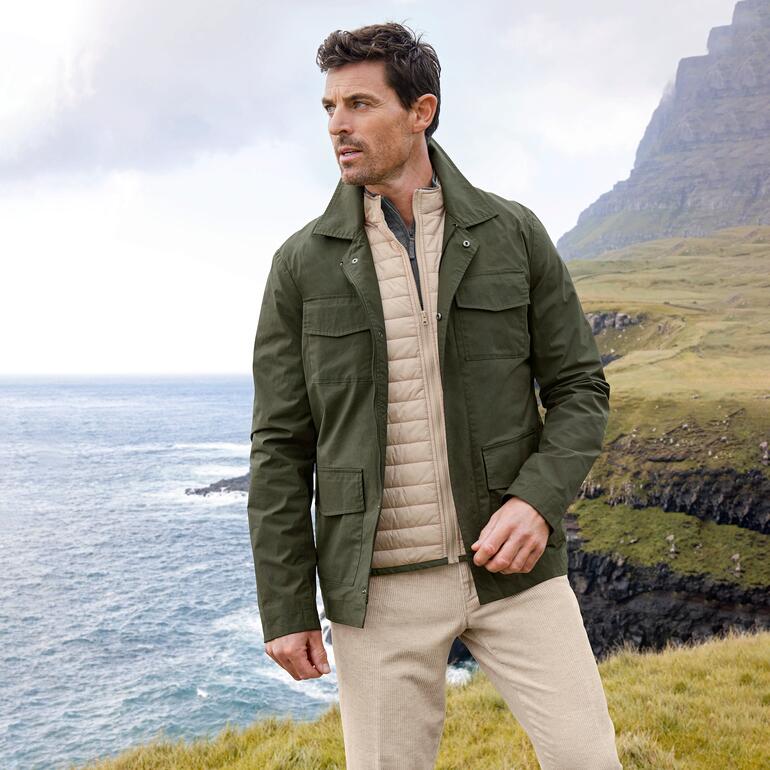 Veste militaire résistante aux intempéries Homme en veste de champ résistante aux intempéries se tenant sur une côte avec une montagne verte en arrière-plan.