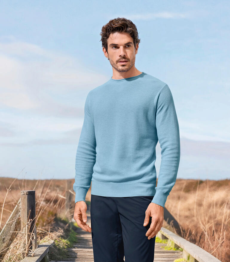 Pull noble en piqué Junghans 1954 Homme dans un pull piqué bleu clair se tenant dans un environnement rural.