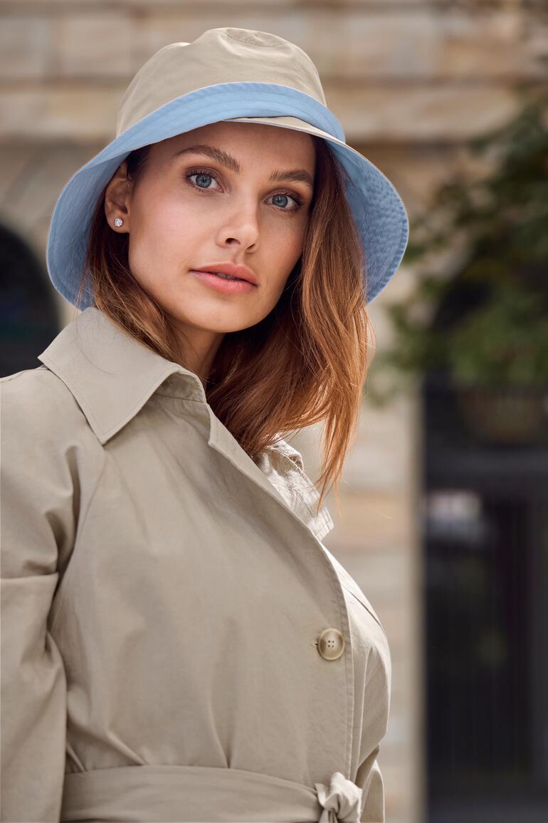 Femme portant un chapeau bucket beige et un trench-coat, regardant la caméra