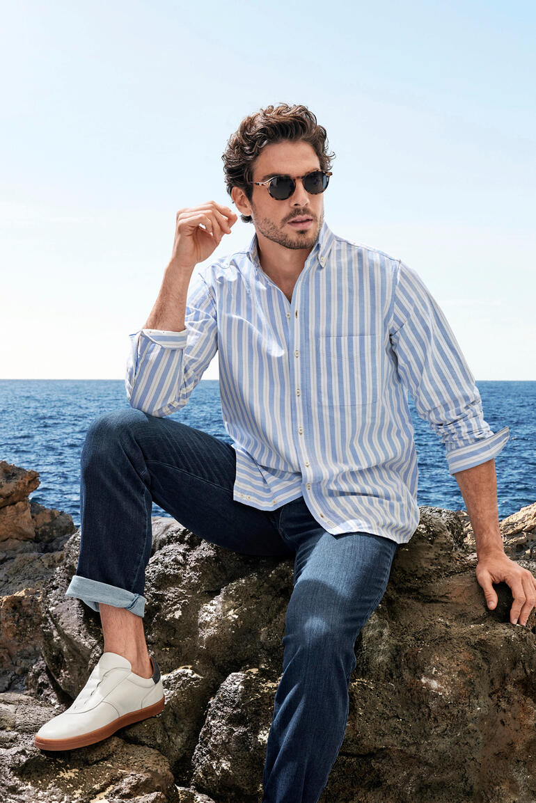 The BDO Shirt, Limited Edition No. 73 Homme en chemise rayée assis sur des rochers au bord de la mer, portant des lunettes de soleil.