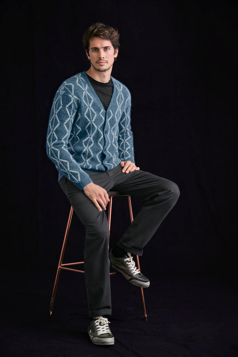 Noir Homme assis sur un tabouret dans un cardigan en alpaga brossé bleu avec un motif losange.
