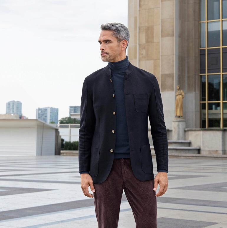Homme en veste décontractée, debout devant un complexe de bâtiments.