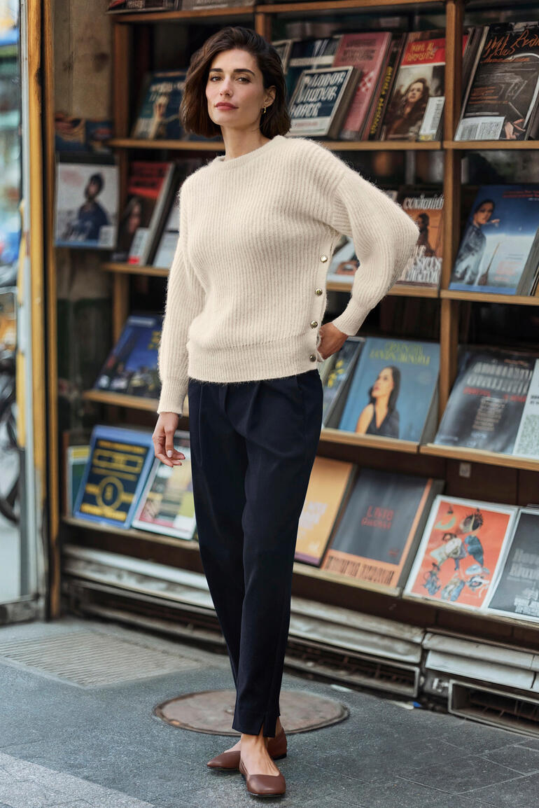 Femme debout devant un kiosque à journaux, portant un pull beige, regardant la caméra.