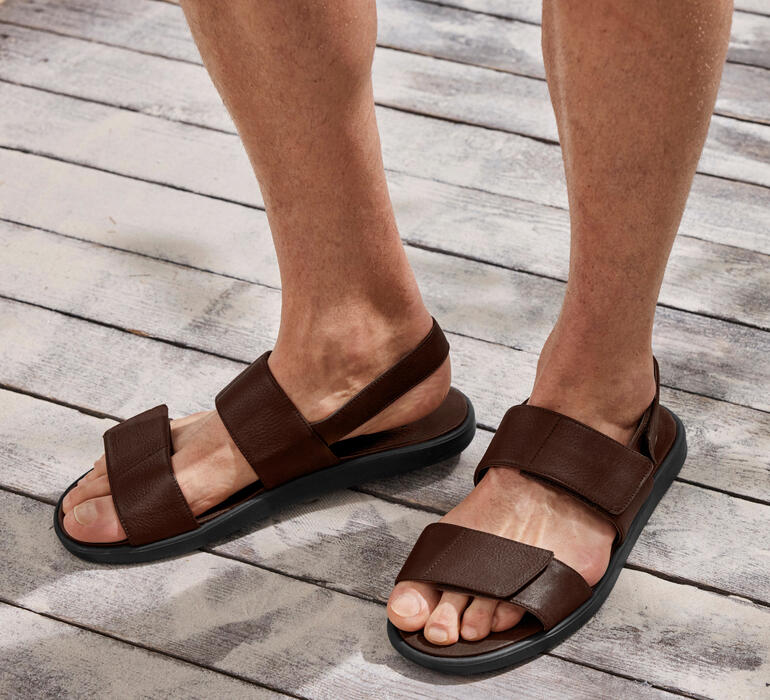 Sandale en cuir brun avec deux lanières sur un pied sur un sol en bois.