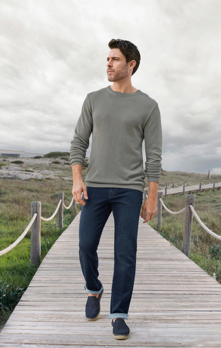 Greige Homme portant un pull Tencel-Piqué Ivanhoe gris et un jean sur une passerelle en bois.