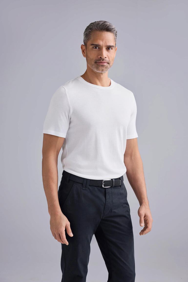 Homme en T-shirt blanc et pantalon noir, se tient devant un fond gris.