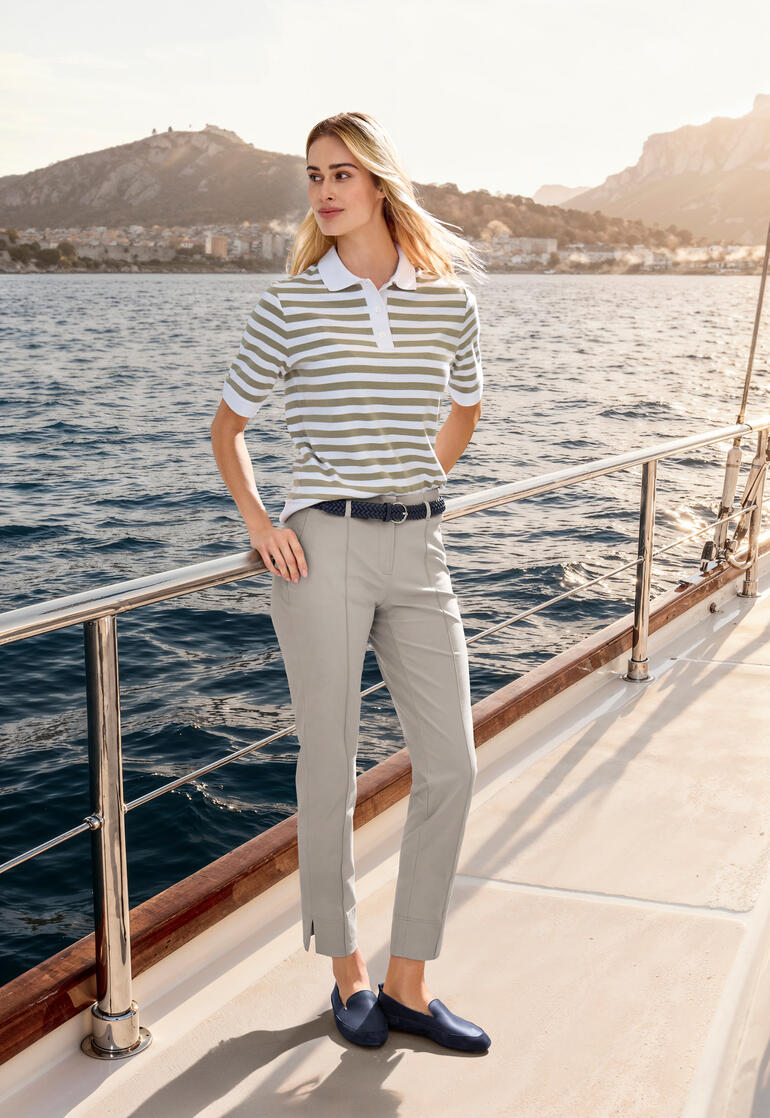 Femme portant un polo boxy beige à rayures, posant au bord d'un lac de montagne.