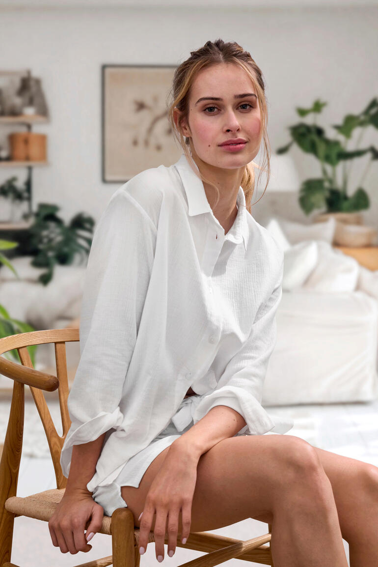 Blouse et Shorts Femme assise sur une chaise, portant une blouse en mousseline blanche Novila, avec des plantes naturelles en arrière-plan.