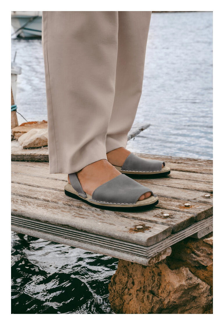 Avarcas de Menorca Personne avec des Avarcas de Menorca grises sur un quai en bois au-dessus de l'eau.
