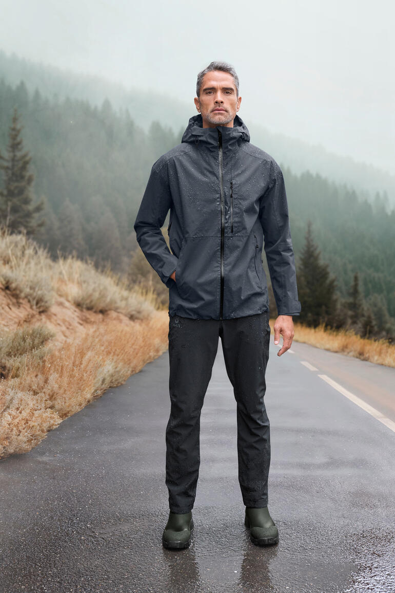 Homme en veste Hardshell grise sur une route mouillée, avec des montagnes en arrière-plan.