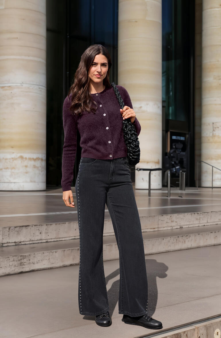 Femme en cardigan bordeaux et pantalon noir devant un bâtiment moderne.