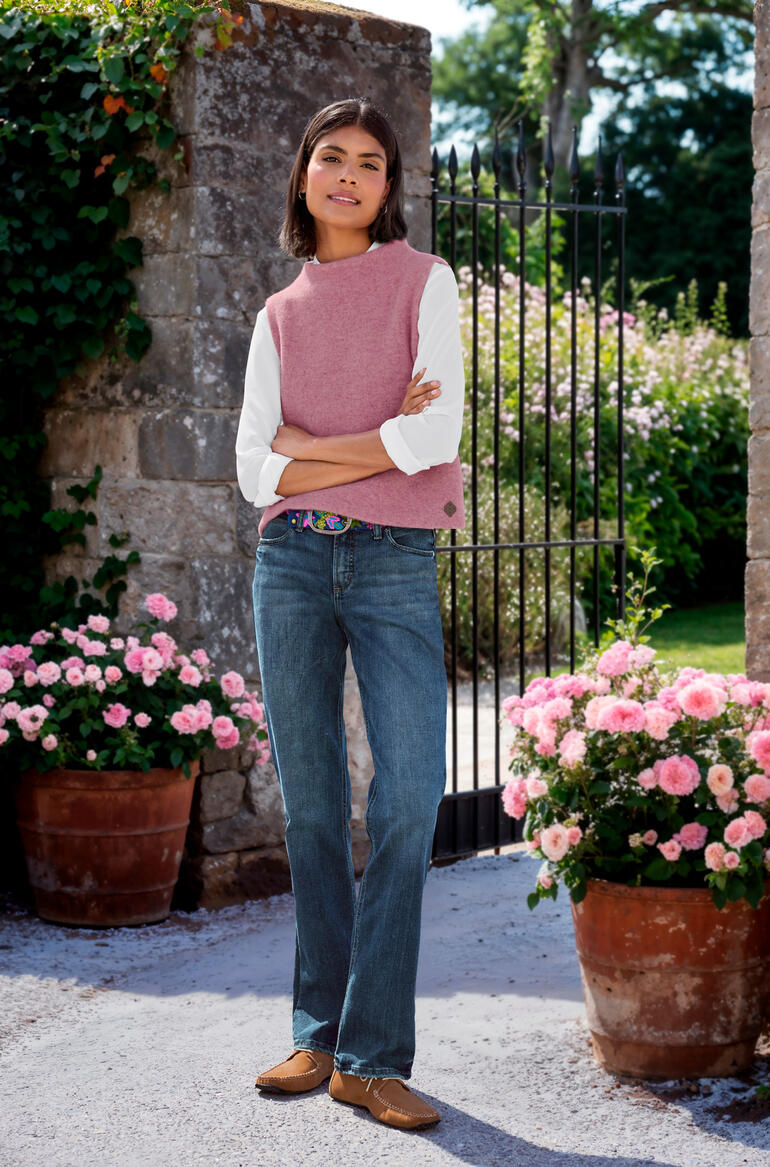 Rose Femme en pull en laine rose sur un t-shirt à manches longues blanc, debout près d'une porte fleurie.