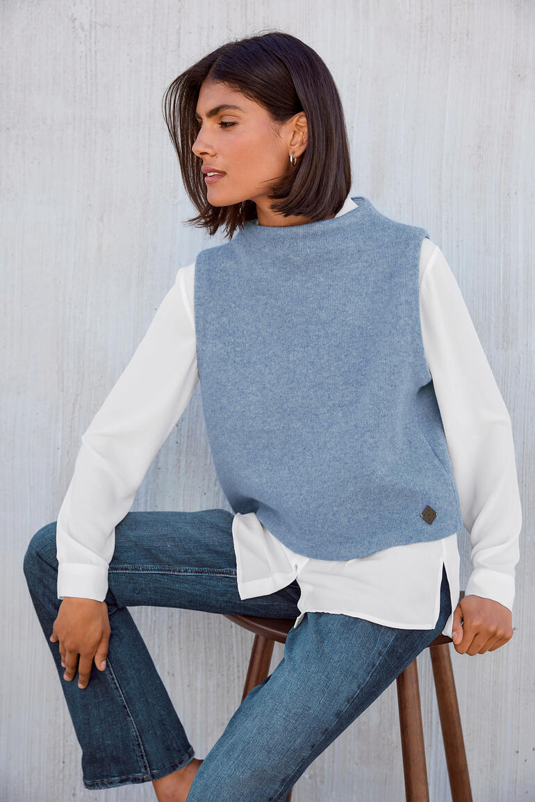 Femme portant un pull en laine bleu sur une chemise blanche, assise sur une chaise.