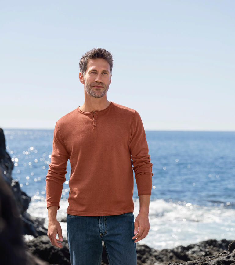 Un homme se tient sur la plage, vêtu d'un pull rouille Ragman et d'un jean.