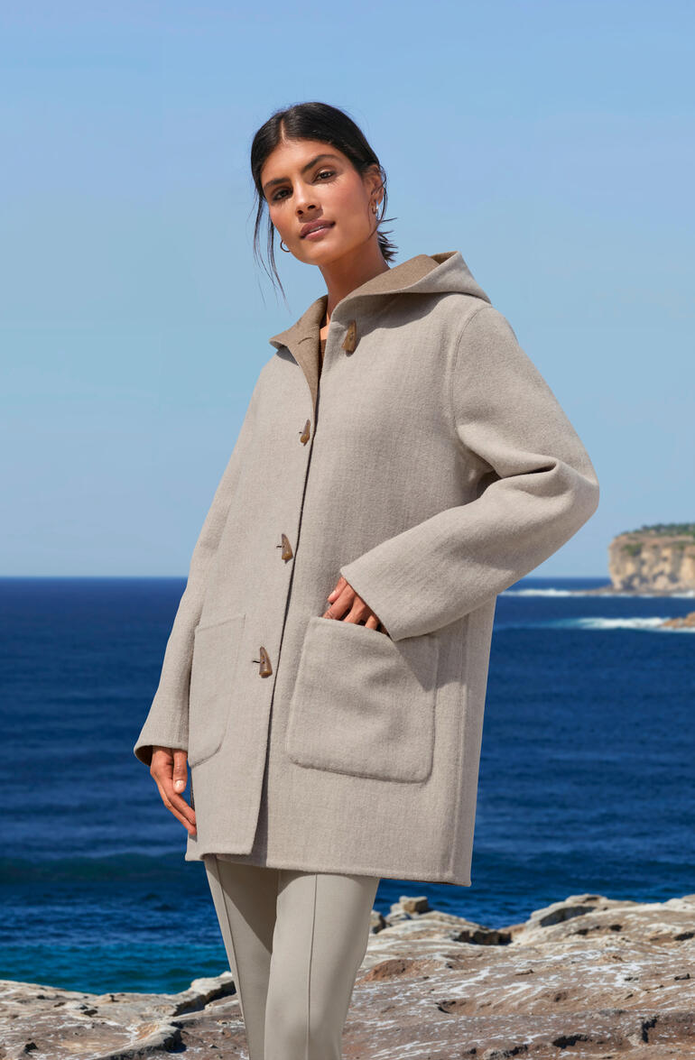 Manteau Duffle à double face en alpaga beige, porté par une femme avec la mer en arrière-plan.