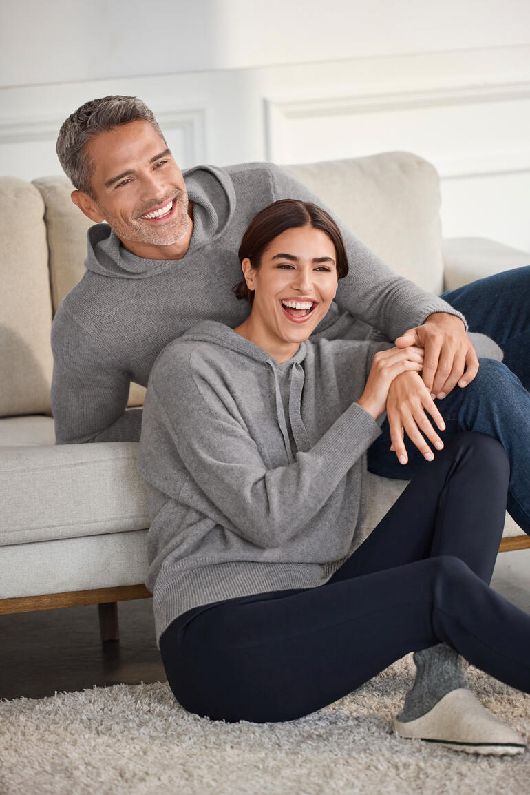 Un couple sourit sur un canapé, portant des sweats à capuche en cachemire bouilli gris