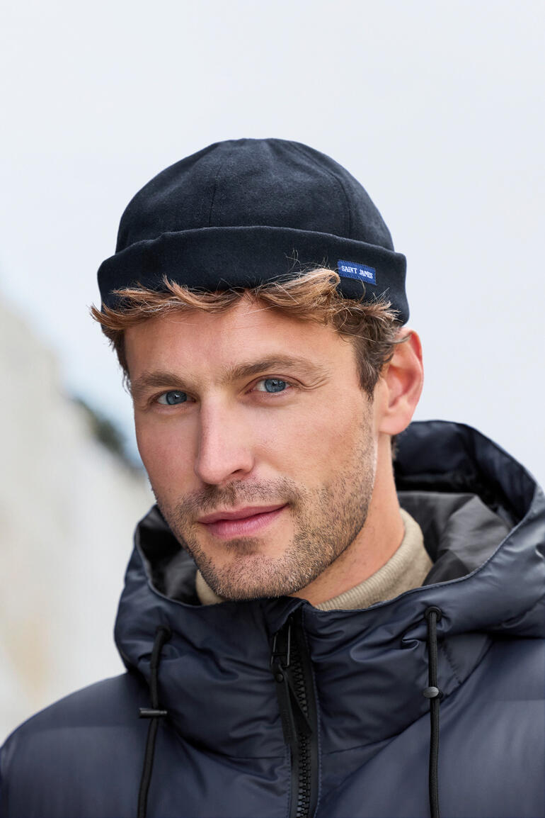 Homme avec un bonnet dans une doudoune, souriant devant un arrière-plan flou.