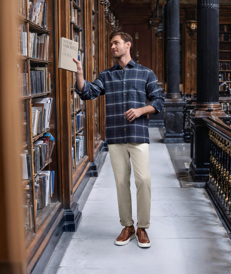 Homme lisant un livre dans une bibliothèque, portant une chemise à carreaux.