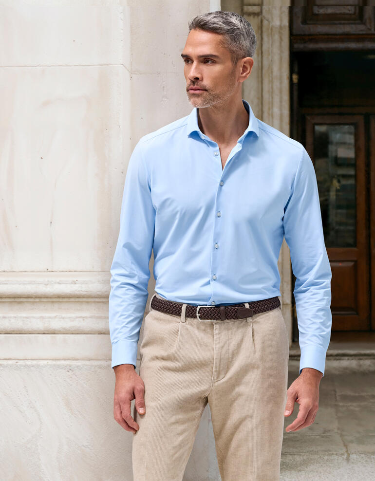 Homme en chemise 24 heures bleu clair, taille 38, devant un fond en pierre.
