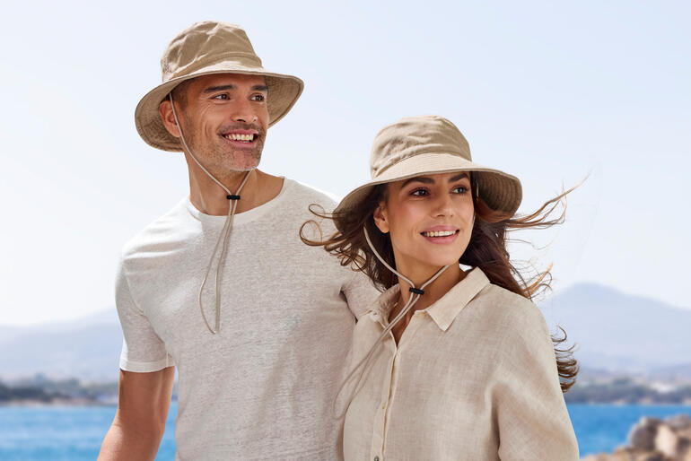 Couple portant des chapeaux de plage couleur sable au bord de la mer, souriant à la caméra.