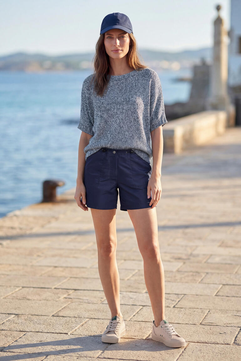 Femme debout face à la mer en pull bleu et short.