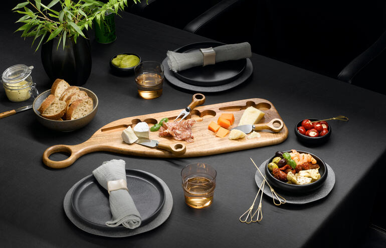 Planche de service design en bois d'acacia avec fromage, fruits et snacks sur une table sombre.