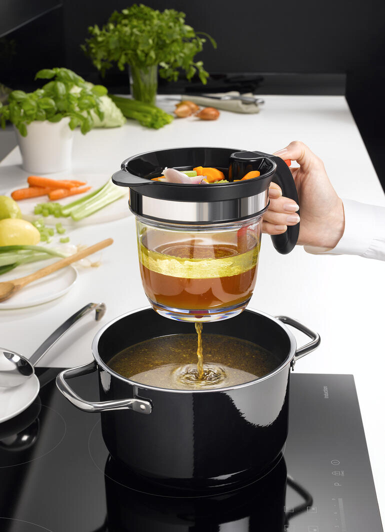 Main utilisant un tamis pour verser du bouillon de légumes dans une casserole, avec une cuisine en arrière-plan.