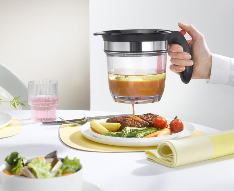 Main avec un tamis versant du bouillon sur une assiette avec des légumes et de la viande, avec une table dressée en arrière-plan.