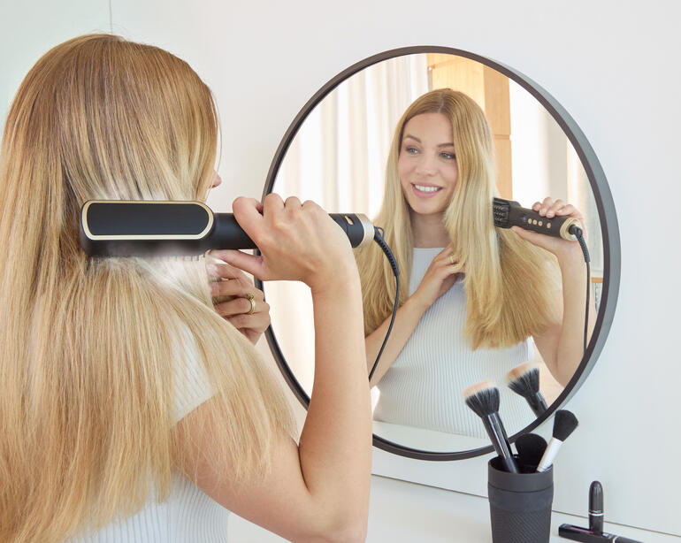 Femme avec une brosse à cheveux ionique devant un miroir dans une pièce moderne.