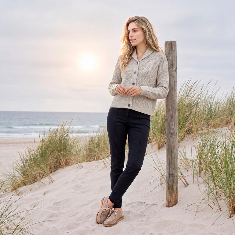Femme en cardigan gris se tenant sur la plage avec le coucher de soleil en arrière-plan.